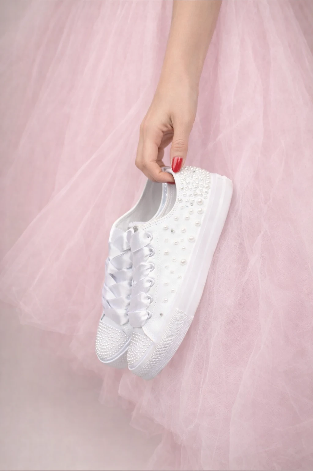 Νύφη με ροζ τούλινο φόρεμα κρατά λευκά Converse sneakers χειροποίητα διακοσμημένα με λευκές πέρλες και σατέν κορδόνια.