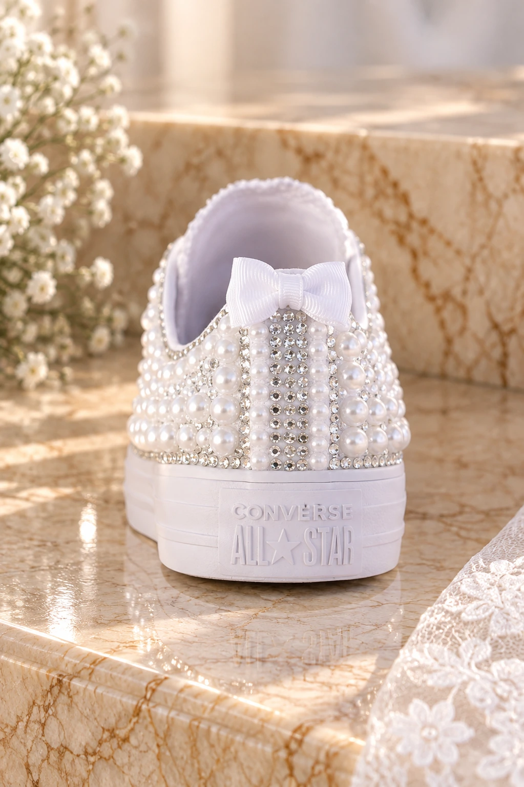 Πίσω όψη λευκού νυφικού Converse, χειροποίητα διακοσμημένου με πέρλες, λαμπερά στρας και λευκό φιόγκο, σε ζεστό μπεζ μαρμάρινο φόντο